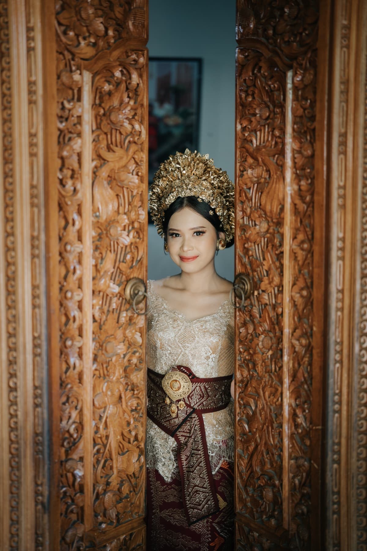 Ima in Balinese wedding dress and gold headpiece, peeking between two carved wooden doors
